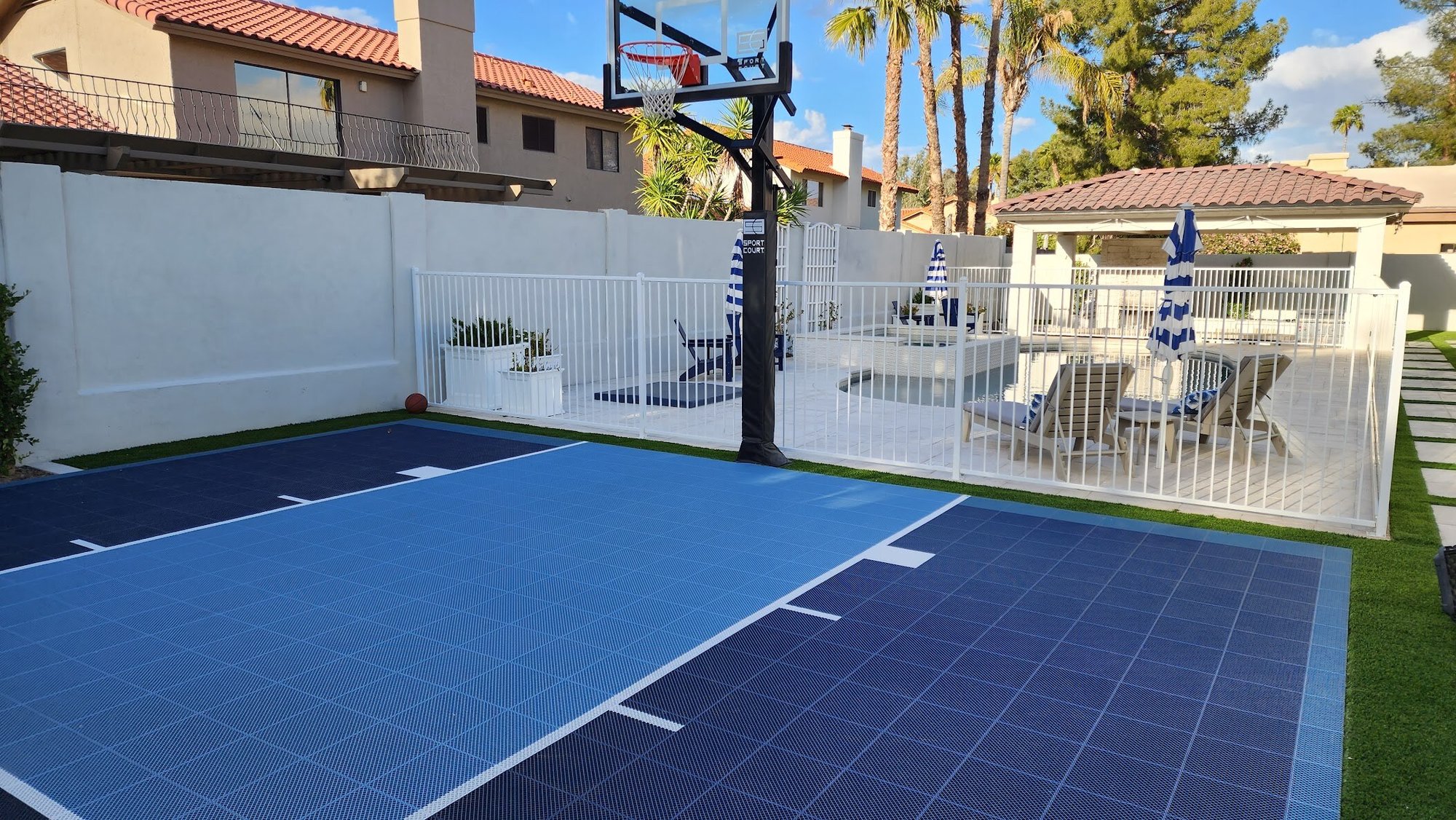 A compact basketball court adjacent to a fenced pool area with seating A compact basketball court adjacent to a fenced pool area with seating